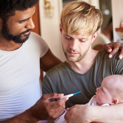 Homokoppel met baby in de armen, twee vaders nemen de temperatuur van hun zieke baby met thermometer