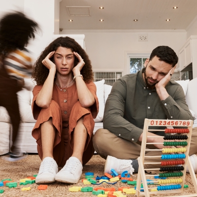 Opvoedstress, ouders zitten uitgeput op de vloer terwijl kinderen rondlopen, speelgoed op de vloer, ouders met opvoedstress