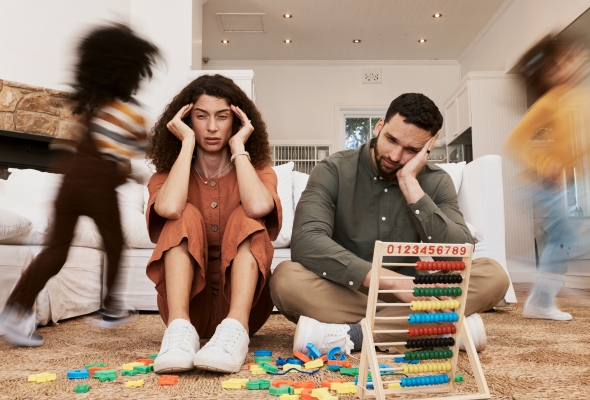 Opvoedstress, ouders zitten uitgeput op de vloer terwijl kinderen rondlopen, speelgoed op de vloer, ouders met opvoedstress
